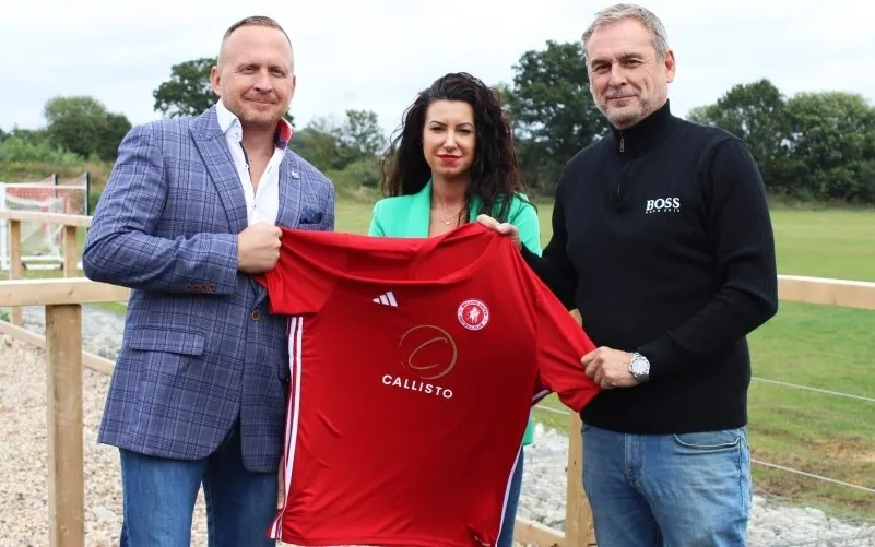 A man, woman, and another man hold up a red Welling United football shirt featuring the Callisto Homes sponsor logo. They are standing pitch-side at a football ground, smiling and marking the announcement of the new club sponsorship. The woman wears a green jacket, while the men wear a check blazer and a black jumper.