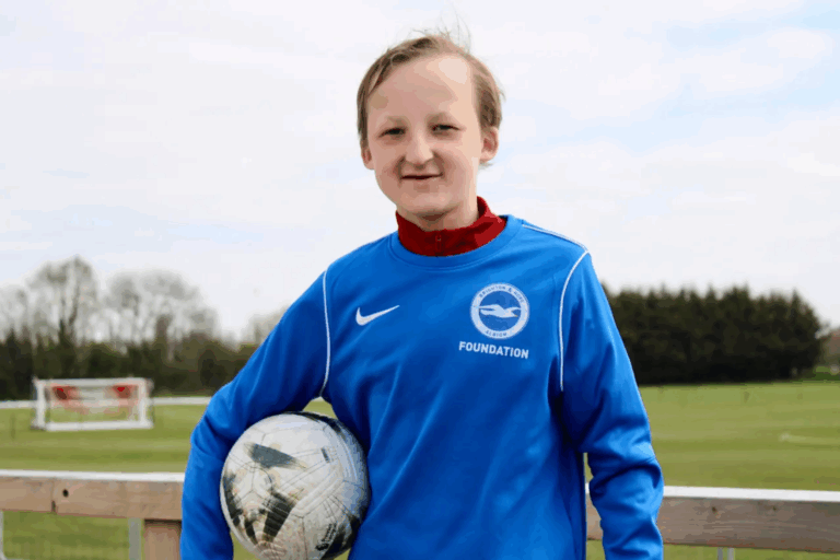Photograph of Thomas Briggs wearing a Brighton and Hove Albion Foundation jumper, holding a football in front of a football pitch