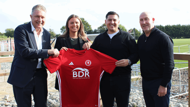 Two men, a woman, and another man stand holding a red Welling United football shirt with the BDR Group sponsor logo. They are smiling and standing on the edge of a football pitch with green fields and goalposts behind them, marking the launch of a new sponsorship deal with BDR.