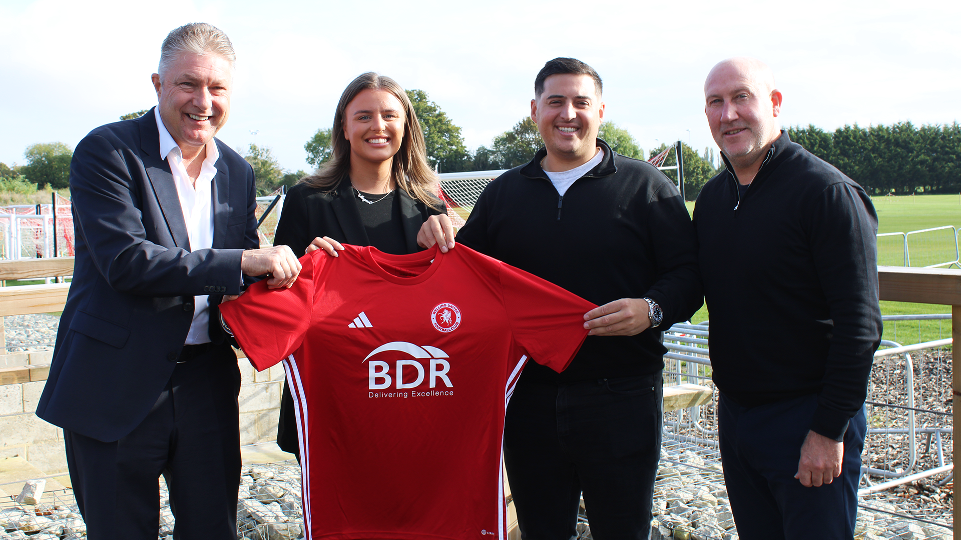 Two men, a woman, and another man stand holding a red Welling United football shirt with the BDR Group sponsor logo. They are smiling and standing on the edge of a football pitch with green fields and goalposts behind them, marking the launch of a new sponsorship deal with BDR.