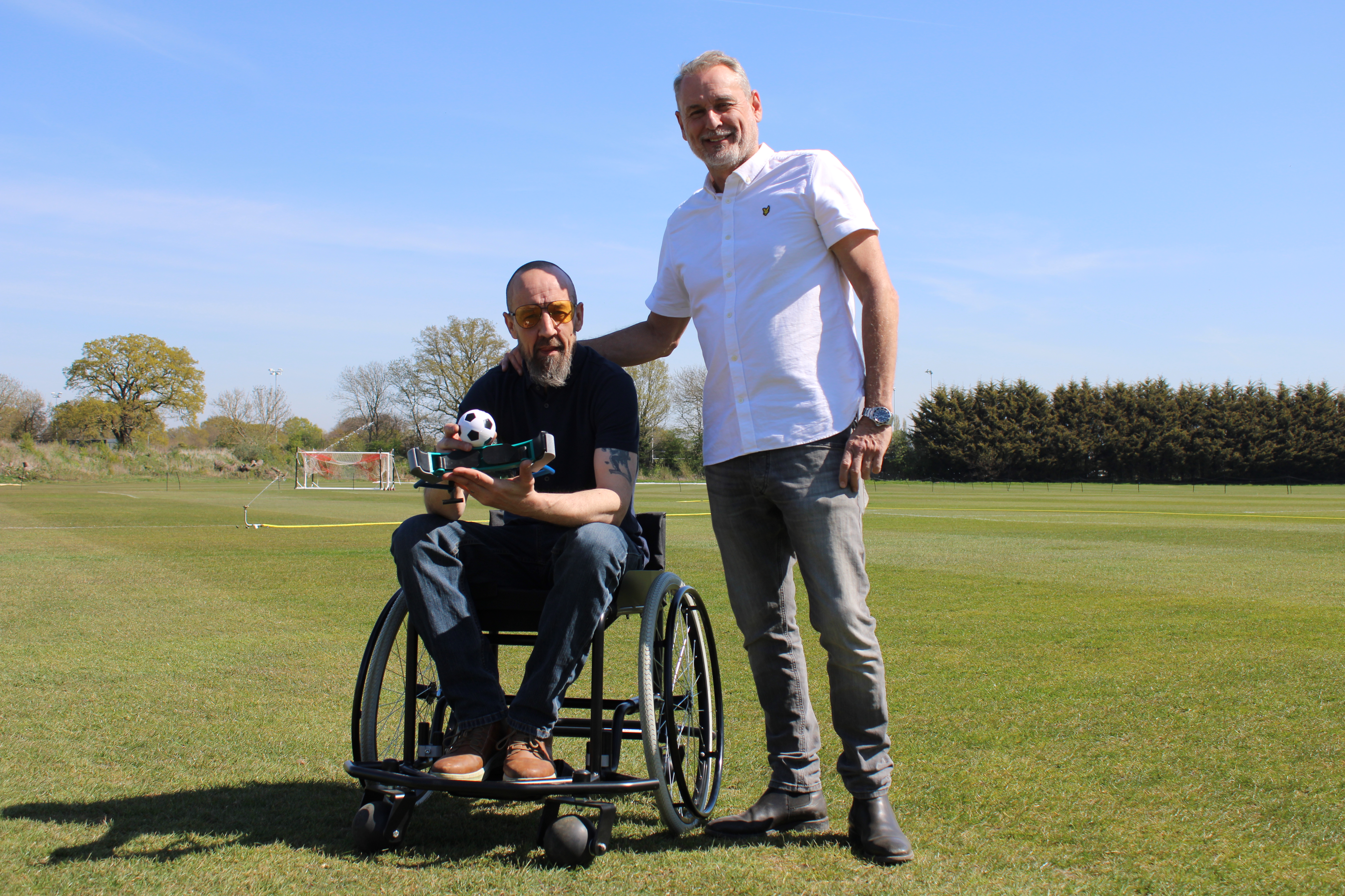 Colin Wood, in a wheelchair, holds a football and prototype applicator on a grass pitch as GB10 Sports Chairman Paul Whitehead stands beside him. The image captures their collaboration to develop wheelchair football for grass pitches through Adaptive Sports Innovations and GB10 Sports.
