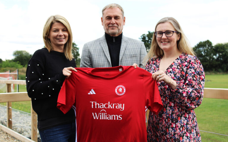 Two women and a man stand together holding a red Welling United football shirt with the Thackray Williams sponsor logo. They are outdoors by the football pitch, smiling at the camera in celebration of the new sponsorship deal.