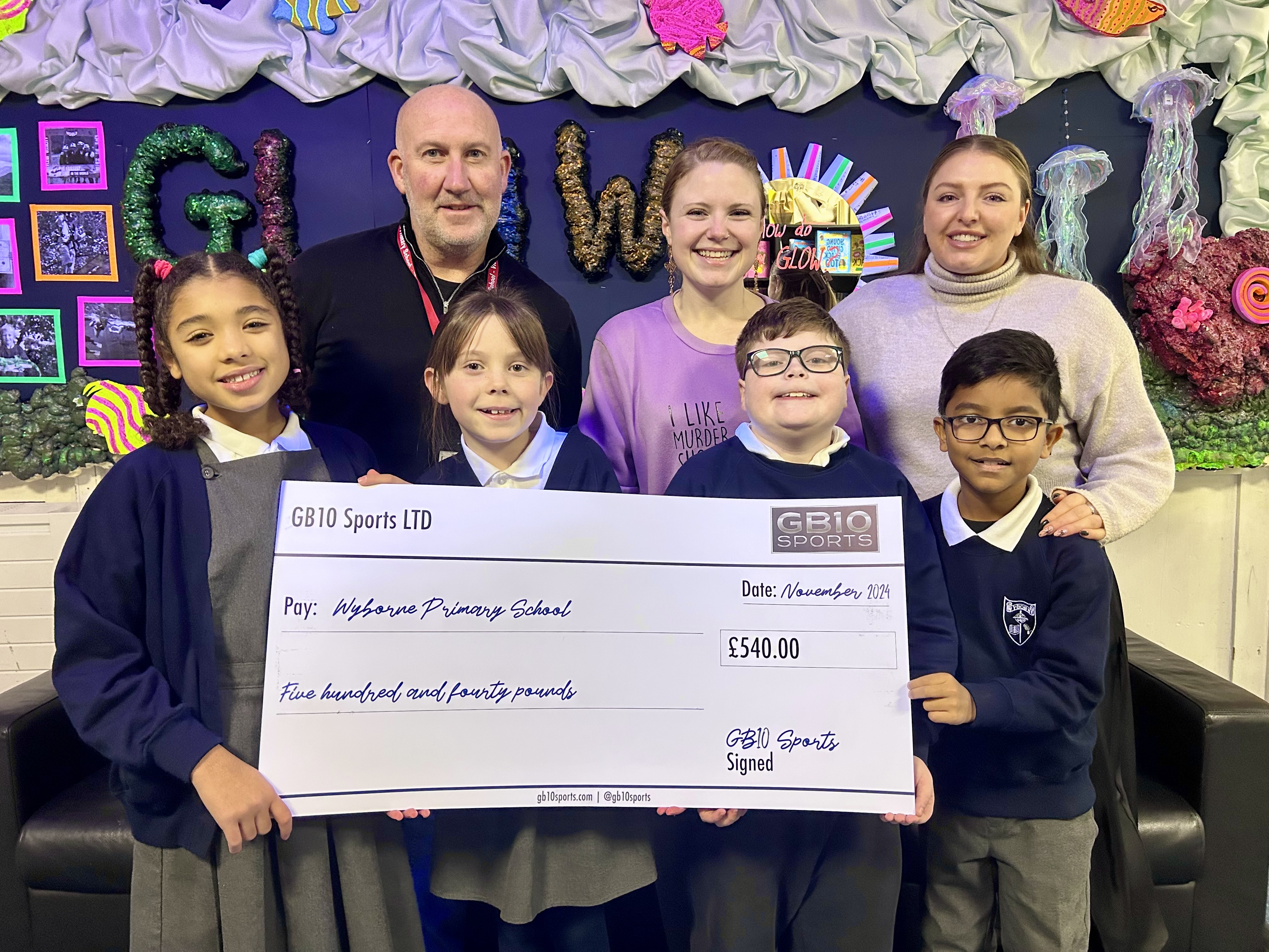 Children and staff from Wyborne Primary School stand with GB10 Sports co-owner Brett Smith, smiling and holding a large cheque for £540, raised during a community fireworks event. The cheque is made out to the school’s PTA and will fund new playground equipment.