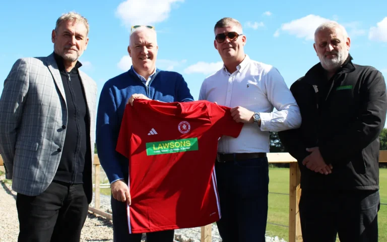 Four men stand pitch-side holding a red Welling United football shirt with the green and yellow Lawsons sponsor logo. The group is gathered to announce a new sponsorship deal, with the shirt held up between the two men in the centre. They are outdoors on a sunny day with a football field behind them.