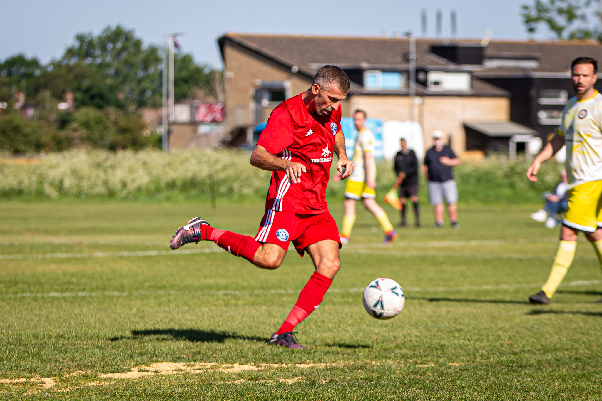 Footballer in a red kit swings his right leg to strike the ball on a sunny grassroots pitch; blurred yellow-kit defenders and clubhouse in background.