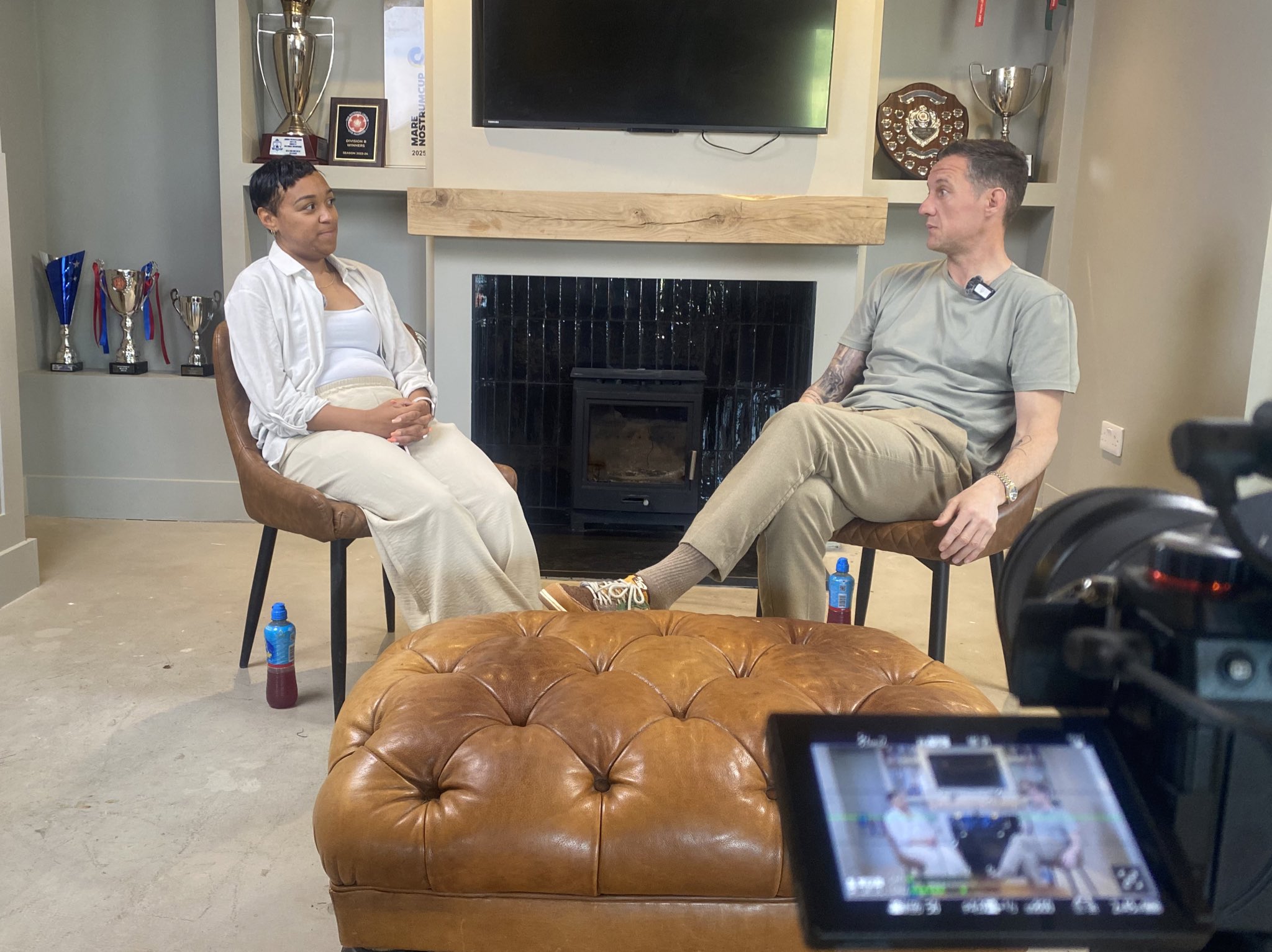 Tony Russell and Sian-Marie film a podcast for urfreshtvsport at GB10, seated in front of AFC Greenwich borough's trophy display in the venue’s media and podcast space.