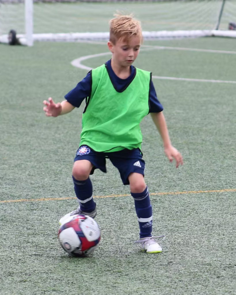 Young footballer in a green bib controlling the ball on a grass football pitch during training