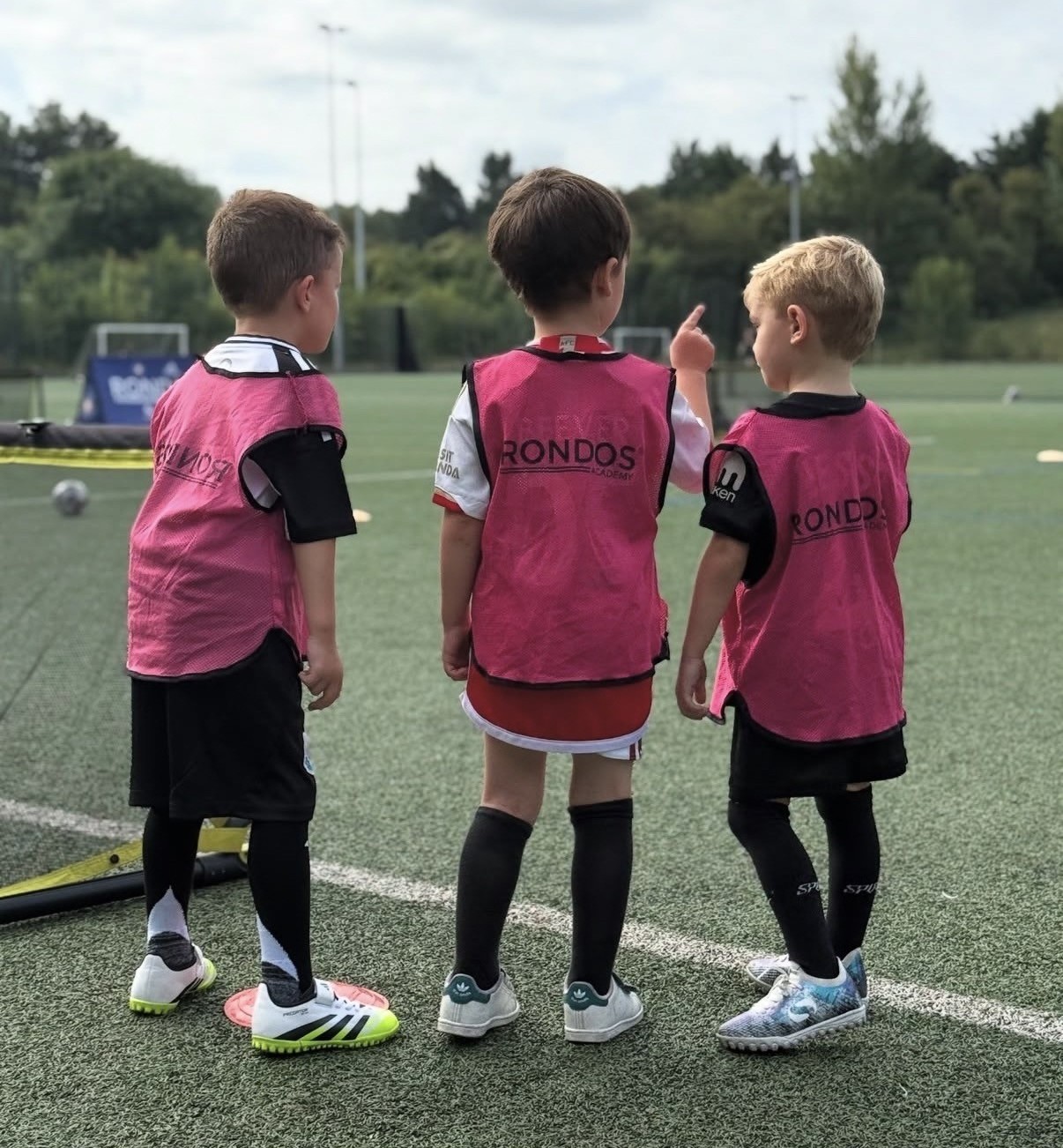 Three young footballers wearing pink bibs stand beside the pitch, discussing the next football training task as the player in the middle points towards the pitch